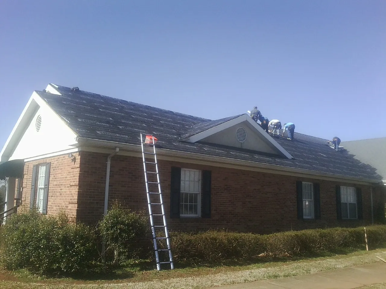 Crew tearing shingles off a brick ranch roof with a ladder set — Metal Roof Repair work by our crew in Caledonia
